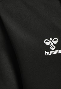 Tessuto atletico nero con una texture liscia, caratterizzato da un logo bianco prominente con un'ape e il testo "hummel". Design semplice e contemporaneo.