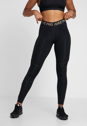 Žena v černých legínách Nike Pro a černých sportovních botách, upravující si pás, pózující proti jednobarevnému světlému pozadí.