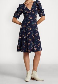Vestido midi floral en color navy con mangas abullonadas, escote en V y falda acampanada. Presenta patrones de flores coloridas con hojas verdes. Lleva botas blancas hasta el tobillo.