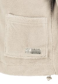 Beige Fleecejacke mit einer Fronttasche, strukturiertem Stoff und einem kleinen Markenetikett. Verfügt über eine Kordel am Saum zur Anpassung.