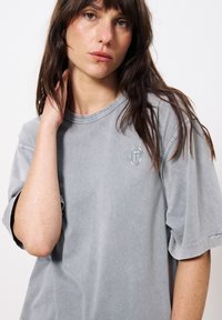 Graues, übergroßes T-Shirt aus weichem Stoff, mit rundem Halsausschnitt und kurzen Ärmeln, sowie einem kleinen gestickten Logo auf der Brust.