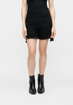 Persoon die zwarte jeansshort met hoge taille en rafelige zomen draagt en zwarte enkellaarzen, staand tegen een effen lichte achtergrond.