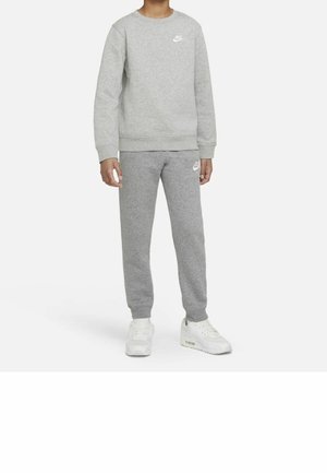 Person iført grå Nike-sweatshirt og matchende sweatpants med hvide Nike-sneakers, stående mod en ensfarvet hvid baggrund.