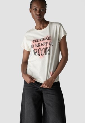 Junge Frau trägt ein weißes T-Shirt mit dem Text "You make my heart go boom!" und schwarze weite Jeans, steht vor einem schlichten grauen Hintergrund.