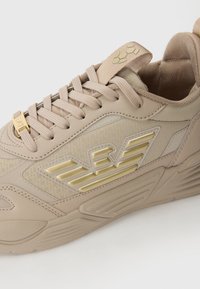 Beige színű sportcipő texturált felülettel, arany díszítéssel és mintás oldallal. Fűszerű záródással és párnázott bokarészzel a kényelem érdekében.