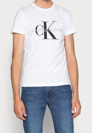 Homme portant un t-shirt blanc Calvin Klein Jeans avec un grand logo « cK » et un jean bleu, montré du cou aux hanches sur un fond uni.