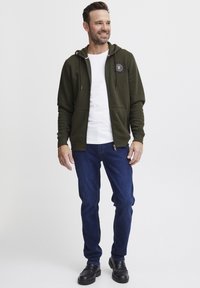 Zelena zip-up dukserica s podešivim špagama, detalj s logotipom, u kombinaciji s bijelom majicom i plavim trapericama. Crne cipele s gumenim potplatima.