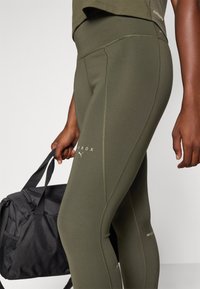 Olivenfarbene Sportleggings aus dehnbarem Stoff mit flachen Nähten. Sichtbares Branding in weißem Text neben dem Saum. Schwarze Sporttasche in der Hand gehalten.