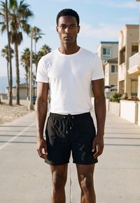 Fehér pólót és fekete rövidnadrágot viselő férfi napfényes tengerparti sétányon áll, háttérben pálmafák és épületek láthatók.
