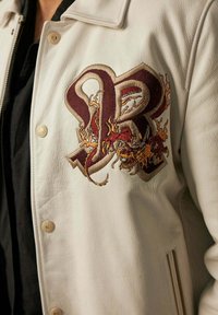 Veste en cuir, couleur crème, avec un design brodé élaboré de dragons et une lettre "R" bordeaux, avec des boutons et une texture visibles.