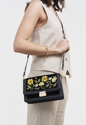 Frau trägt beige ärmelloses Oberteil und Hose, hält schwarze Umhängetasche mit gelber Blumenstickerei und goldenem Verschluss.