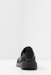 Zapatilla deportiva negra con parte superior de punto texturizado, acentos dorados y suela de patrón multicolor. Tacón redondeado y tirador en la parte posterior.
