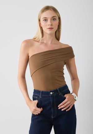 Bodysuit marron décolleté en tissu doux et extensible, associé à un jean en denim bleu foncé taille haute. Le modèle pose les mains sur les hanches.