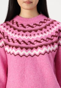 Rosa stickad tröja med ett mönstrat yoke med vita och mörkbruna zigzag- och vågmotiv. Texturen verkar mjuk och kraftig.