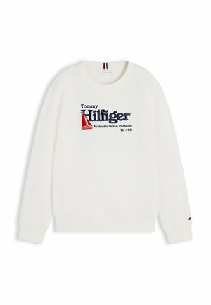 Junge steht mit den Händen in den Taschen, trägt ein weißes Tommy Hilfiger Sweatshirt, blaue Jeans und weiße Sneakers vor weißem Hintergrund.