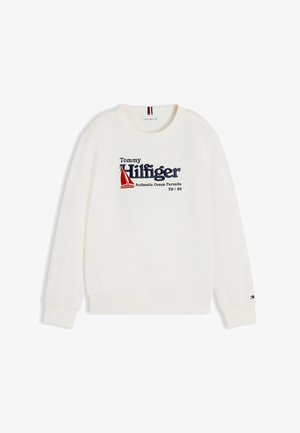 Jongen staat met zijn handen in zijn zakken, draagt een wit Tommy Hilfiger sweatshirt, blauwe spijkerbroek en witte sneakers tegen een witte achtergrond.