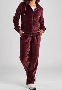 Burgundský velurový teplákový kompletní s zipovou mikinou, předními kapsami a odpovídajícími kalhotami. Obsahuje elastické manžety a pas. Stylizováno se sneakers.