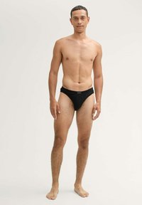 Fekete bikini stílusú fürdőruha, puha anyagból; alacsony derékú és minimális takarást biztosító kialakítással. A modell függőlegesen állva, mezítláb pózol.