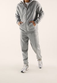 Hombre vestido con una sudadera con cremallera de color gris claro y pantalones de chándal a juego, llevando unas zapatillas de correr blancas y plateadas, de pie sobre un fondo neutro.