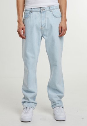 Světle modré džíny s rovným střihem nošené osobou, která stojí, v kombinaci s bílými teniskami a zastrčenou bílou košilí.