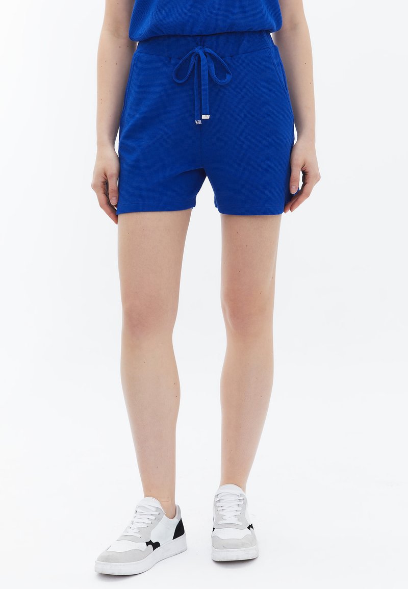 Blauwe gebreide shorts met een elastische tailleband en koord. Heeft een textuurstof en een relaxte pasvorm, gecombineerd met witte sneakers.