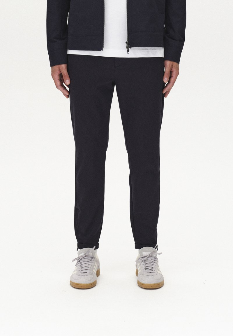 Mornarske jogger hlače s slim fit krojem, ki imajo zadrge na gležnjih in gladko teksturo, v kombinaciji z svetlosivimi semiš športnimi čevlji.