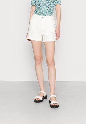 Personne portant un short blanc, un haut fleuri bleu-vert et des sandales blanches à semelles noires, debout sur un sol gris contre un mur blanc.