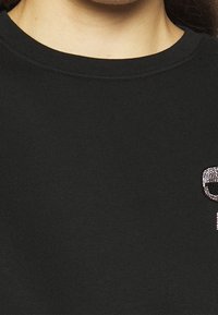 Svart sweatshirt med rund halsringning, med ett strukturerat mönster och strassdekorationer i ett mönster på vänstra bröstområdet.