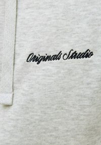 Tissu gris avec texte brodé noir indiquant "Originals Studio" près d'un cordon tissé sur le côté gauche.