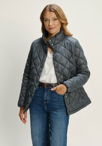 Žena v šedej prešívanej bundě, bílem tričku s výstřihem do V, modrých džínách a hnědém pásku, stojí s jednou rukou v kapse.