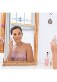 Facial cleansing device in white; user applying it to her cheek. Bamboo-framed mirror and skincare products in soft pink tones visible.