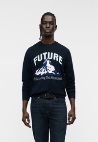 Ναυτικό πουλόβερ με τη λέξη «FUTURE» και γραφικό βουνό σε λευκό και μπλε. Φοριέται με σκούρο τζιν. Απλός στρογγυλός λαιμός και ριμπ μανσέτες.