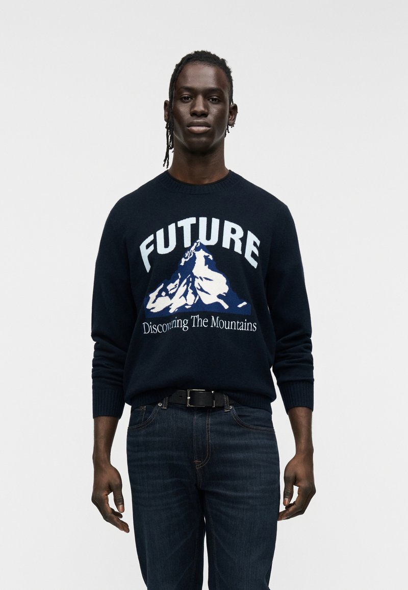 Ναυτικό πουλόβερ με τη λέξη «FUTURE» και γραφικό βουνό σε λευκό και μπλε. Φοριέται με σκούρο τζιν. Απλός στρογγυλός λαιμός και ριμπ μανσέτες.