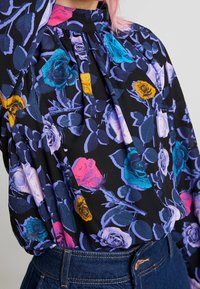 Blouse noire avec un motif floral coloré comprenant des roses roses, bleues et jaunes. Le tissu a une texture douce et se fronce au niveau de l'encolure.
