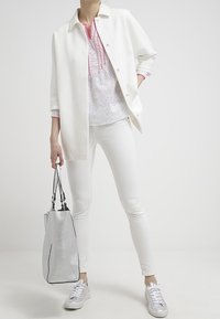 Weiße oversized Jacke mit Knöpfen, über einem weißen Oberteil mit rosa Akzenten getragen. Weiße Skinny Jeans und eine silberne, texturierte Handtasche vervollständigen den Look.