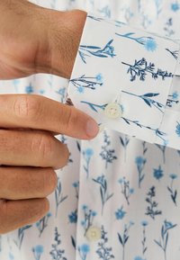 Băgarea unui nasture în manșeta unei cămăși albe cu un model floral albastru, arătând un prim-plan al degetelor și detaliilor textile.