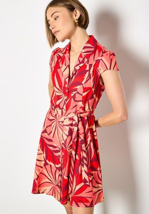 Femme aux cheveux courts portant une robe à manches courtes rouge et rose à motif de feuilles, avec des boutons et une ceinture à la taille, debout devant un fond blanc.