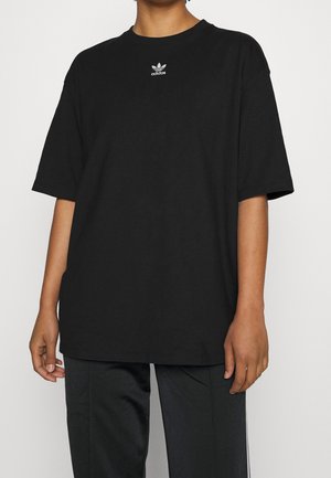 Persoon die een losse zwarte Adidas T-shirt draagt met een klein wit logo op de borst en een zwarte broek tegen een effen achtergrond.