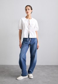 Witte katoenen blouse met pofmouwen en een strikdetail aan de voorkant, gecombineerd met losse blauwe denim jeans en witte sneakers met gouden accenten.