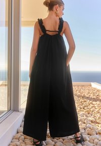 Crna maxi haljina s volanastim naramenicama, otvorenim leđima i ležernim krojem. Glatka tkanina elegantno pada. Crne sandale upotpunjuju izgled.