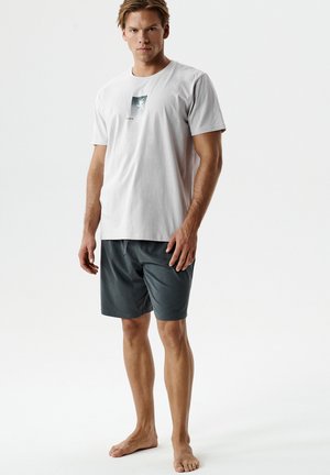 Mężczyzna stojący boso, ubrany w jasnoszarą koszulkę z nadrukiem i ciemnoszare krótkie spodenki do kolan na jednolitym białym tle.