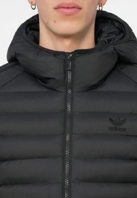 Melnā pūkām vāciņš ar rāvējslēdzēja priekšpusi, kapuci un horizontāli quiltētu tekstūru. Iespējots reljefs Adidas logo uz krūtīm.