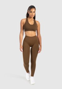 Donna in piedi con indosso un reggiseno sportivo seamless marrone e leggings a vita alta con sneakers bianche su sfondo neutro.