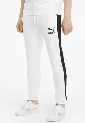 Osoba nosząca białe spodnie dresowe Puma z czarnymi paskami po bokach oraz białe sneakersy Puma, stojąca na tle jednolitego białego tła.