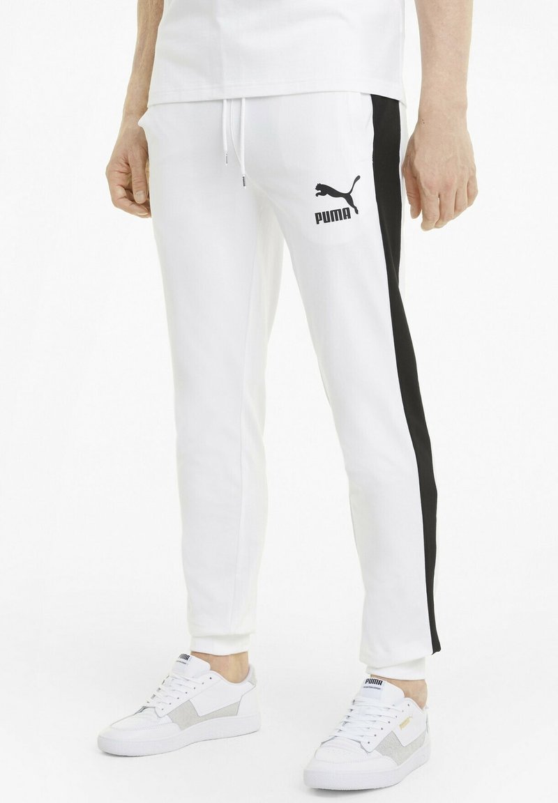Personne portant un pantalon de survêtement blanc Puma avec des bandes noires sur les côtés et des baskets blanches Puma, debout devant un fond blanc uni.