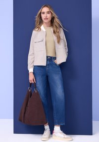 Femme portant une veste beige et un haut kaki, jean bleu, baskets blanches, tenant un grand sac à main marron devant un mur bleu.