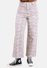 Jean à motif floral dans des couleurs pastel avec une taille haute, une coupe jambes larges, et une étiquette sur la poche avant. Porté avec des baskets montantes noires.
