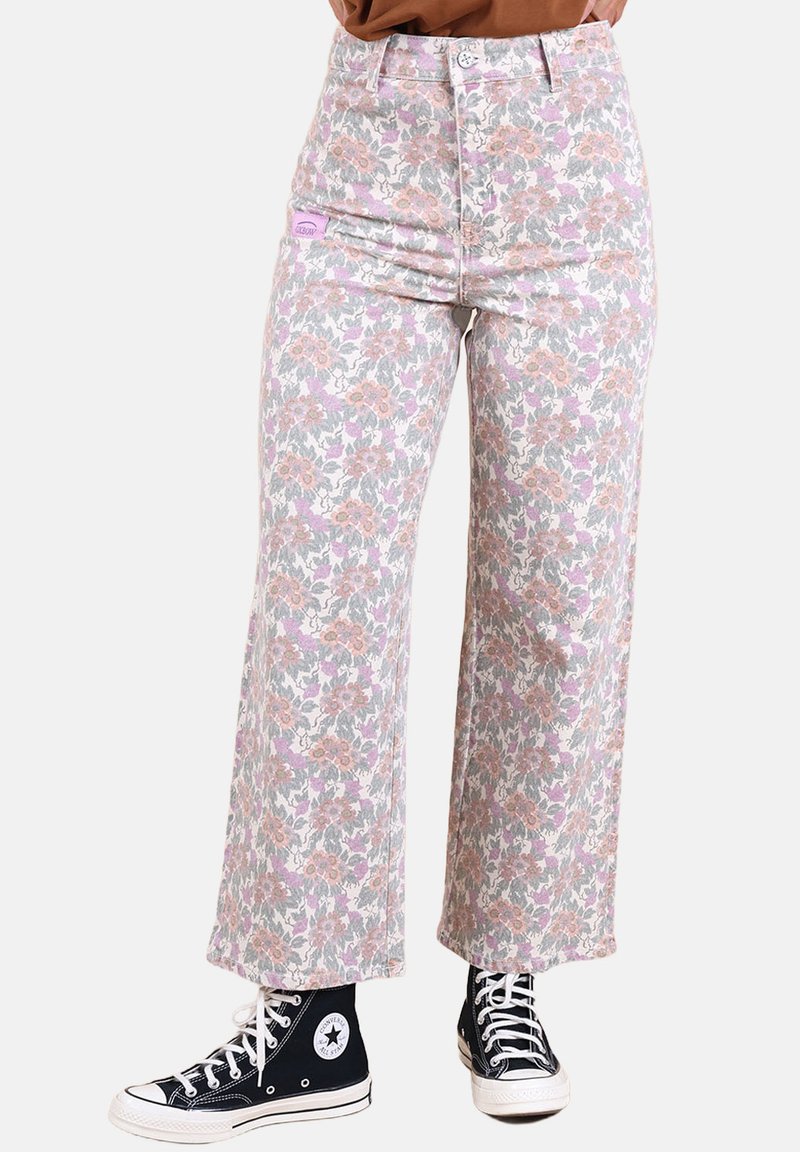 Jean à motif floral dans des couleurs pastel avec une taille haute, une coupe jambes larges, et une étiquette sur la poche avant. Porté avec des baskets montantes noires.