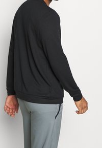 Sudadera negra de manga larga y cuello redondo, hecha de una tela suave. Presenta un corte relajado y un dobladillo acanalado, combinada con pantalones grises.