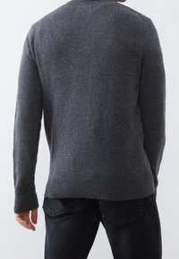 Grauer Strickpullover mit rundem Halsausschnitt und geripptem Saum, kombiniert mit schwarzer Jeans. Die Textur wirkt weich und glatt.